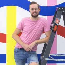 Spencer is leaning against a ladder with a colourful wall mural behind him